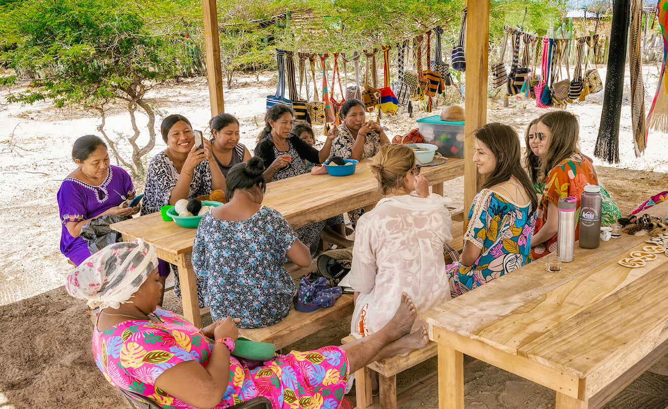 Group of women sitting around wooden tables outdoors, engaged in conversation.